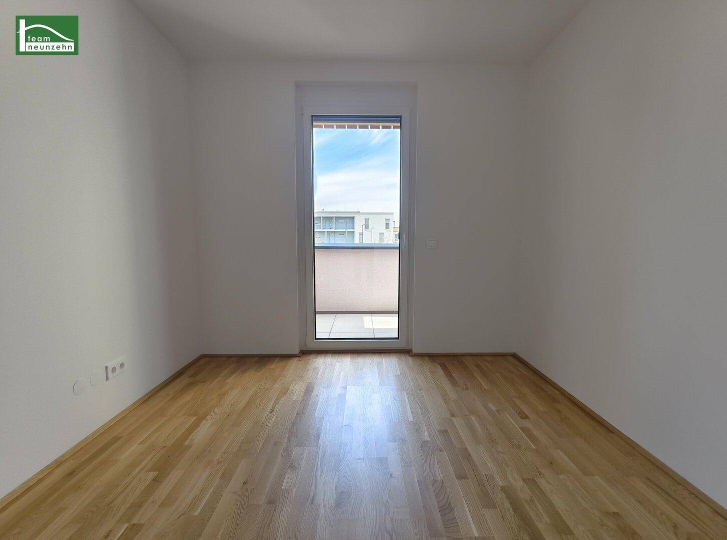 Leeres Zimmer mit Parkettboden und einer Tür, die zu einem Balkon führt.