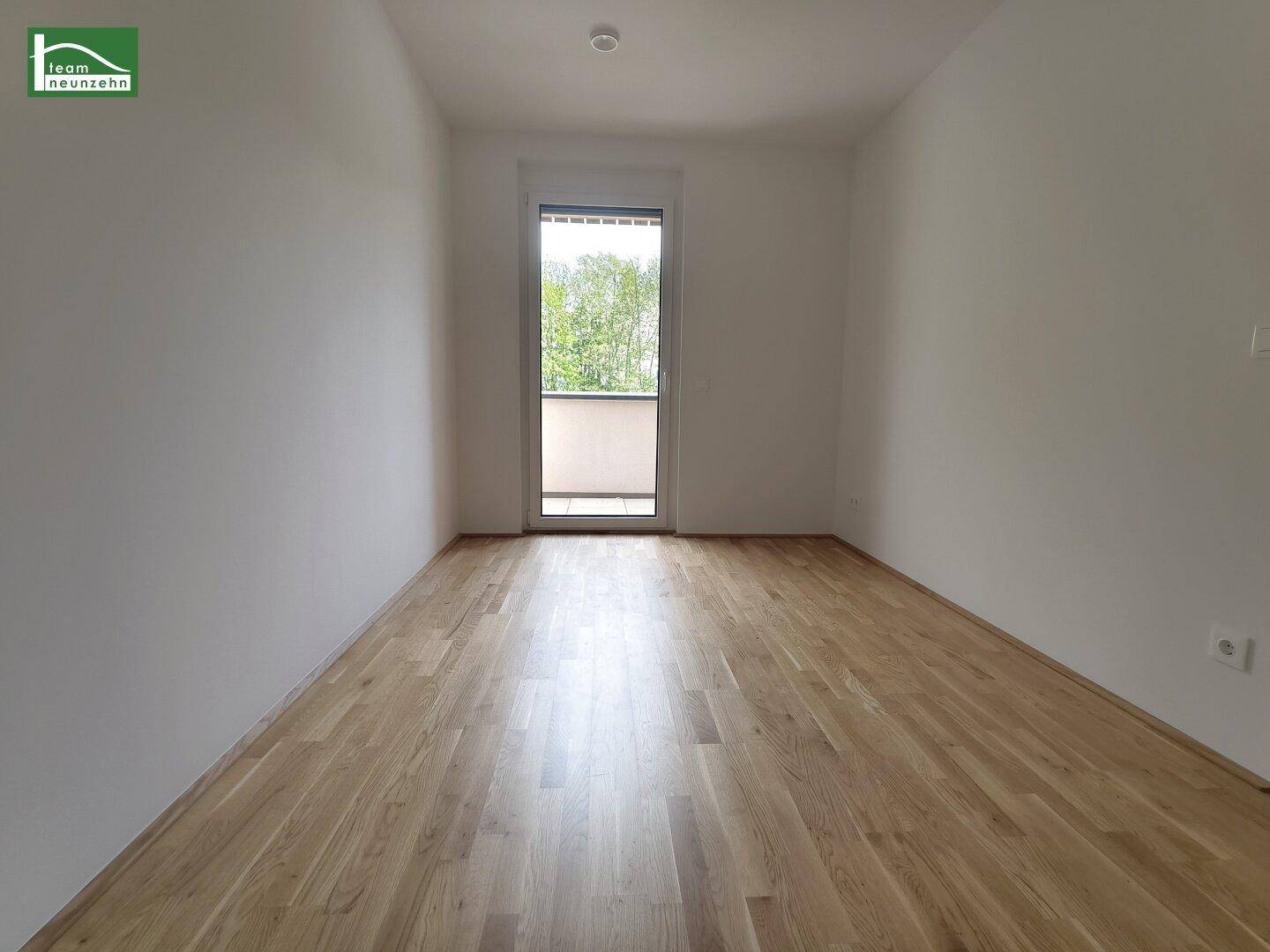 Leeres Zimmer mit Parkettboden und einer Tür, die zu einem Balkon führt.