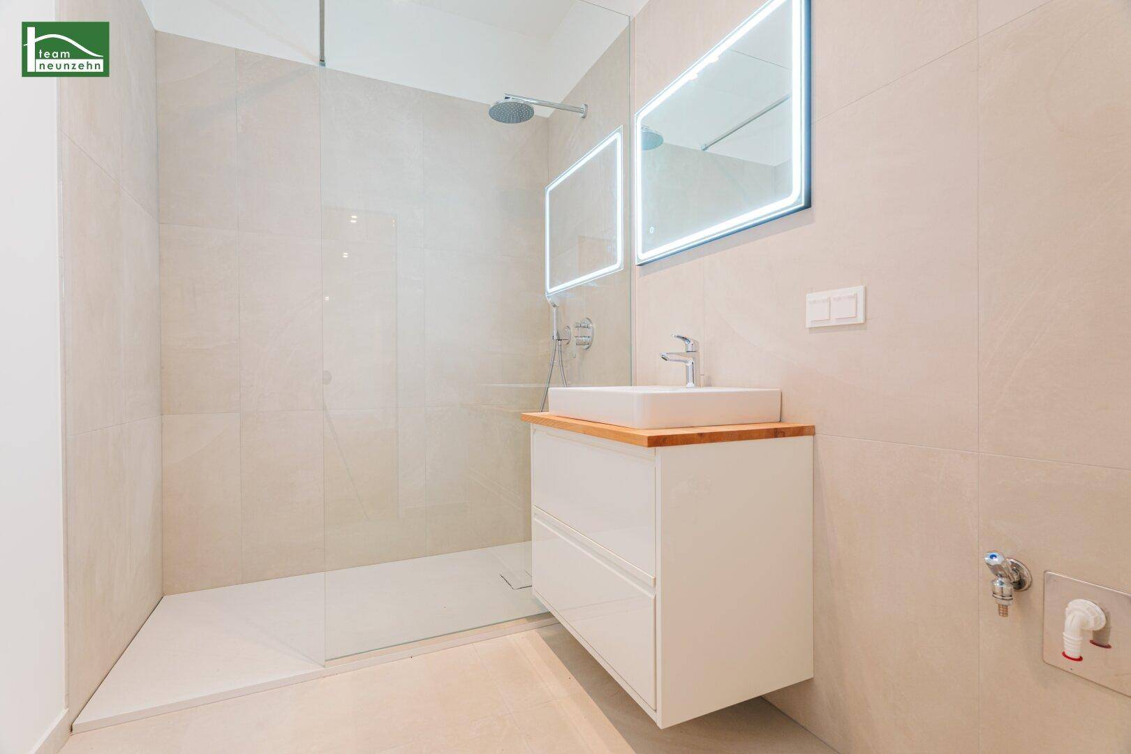 Modernes Badezimmer mit ebenerdiger Dusche, Waschtisch und beleuchtetem Spiegel, in hellem Design.