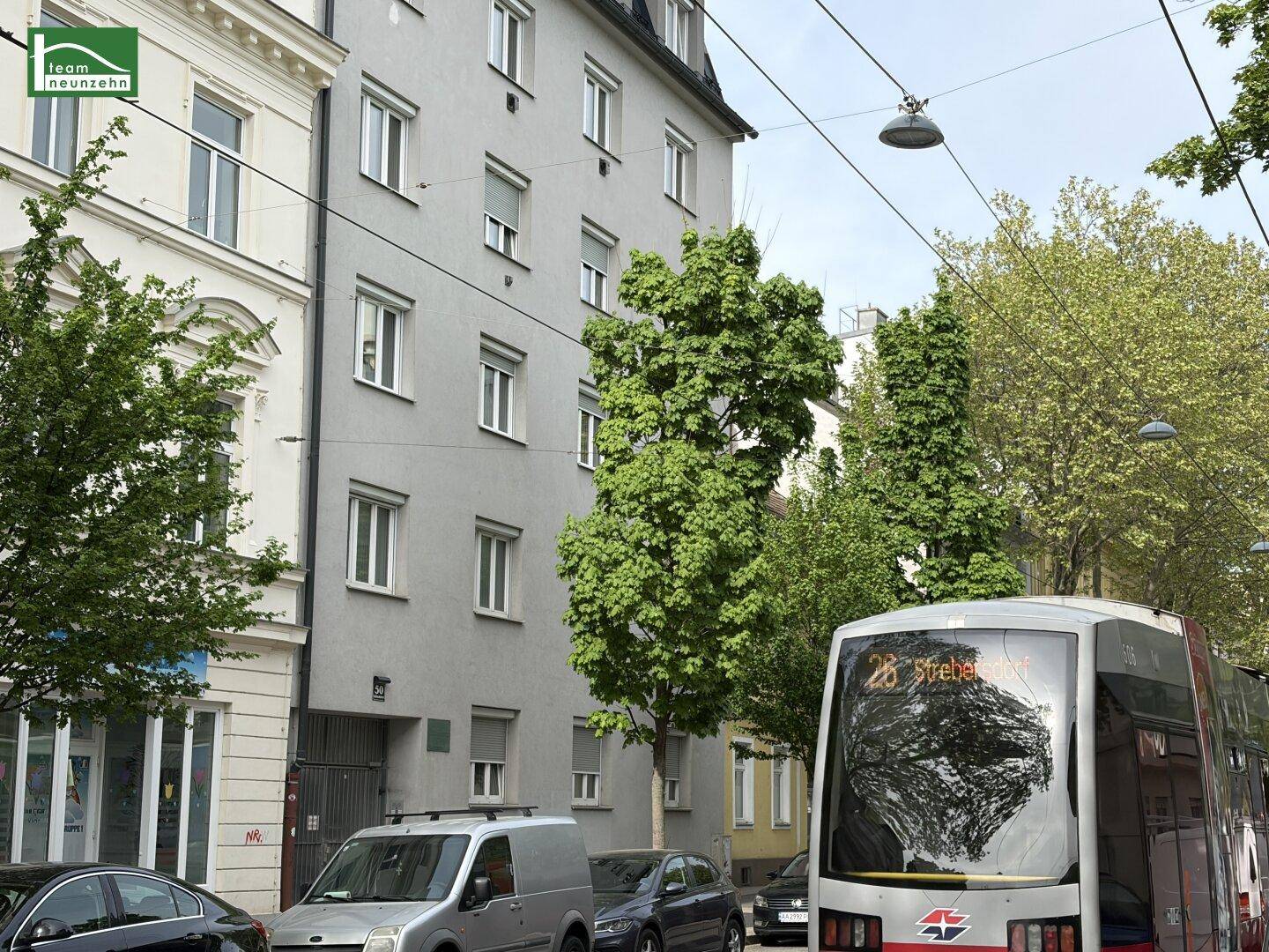 Straßenansicht des Wohngebäudes mit parkenden Autos und einer vorbeifahrenden Straßenbahn.