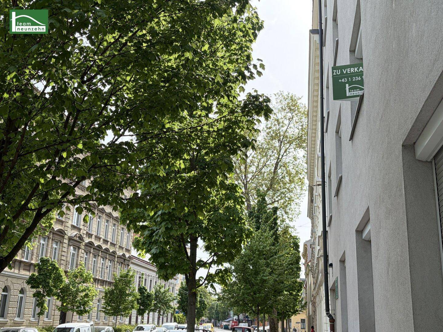 Baumgesäumte Straße mit historischen Gebäuden und einem 'Zu Verkaufen'-Schild an der Fassade.