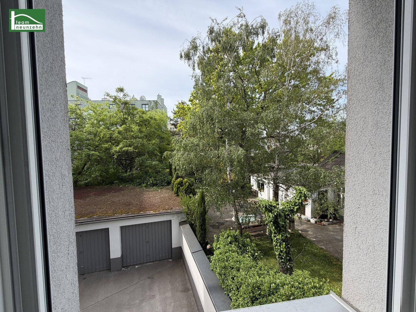 Ausblick aus dem Fenster auf einen grünen Innenhof mit Garagen und hohen Bäumen.