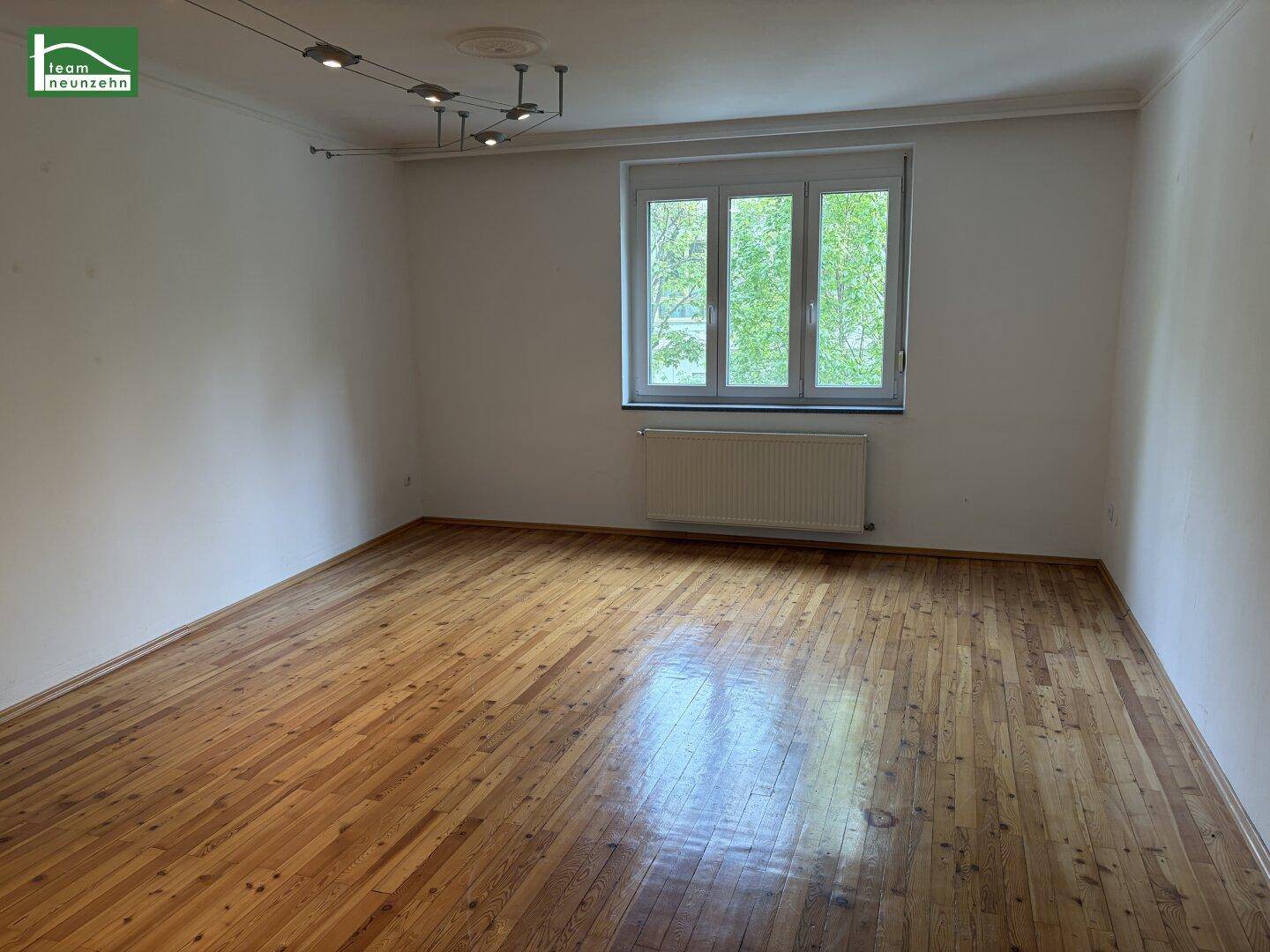 Helles Wohnzimmer mit glänzendem Holzparkettboden und einem großen Fenster zur Straße.