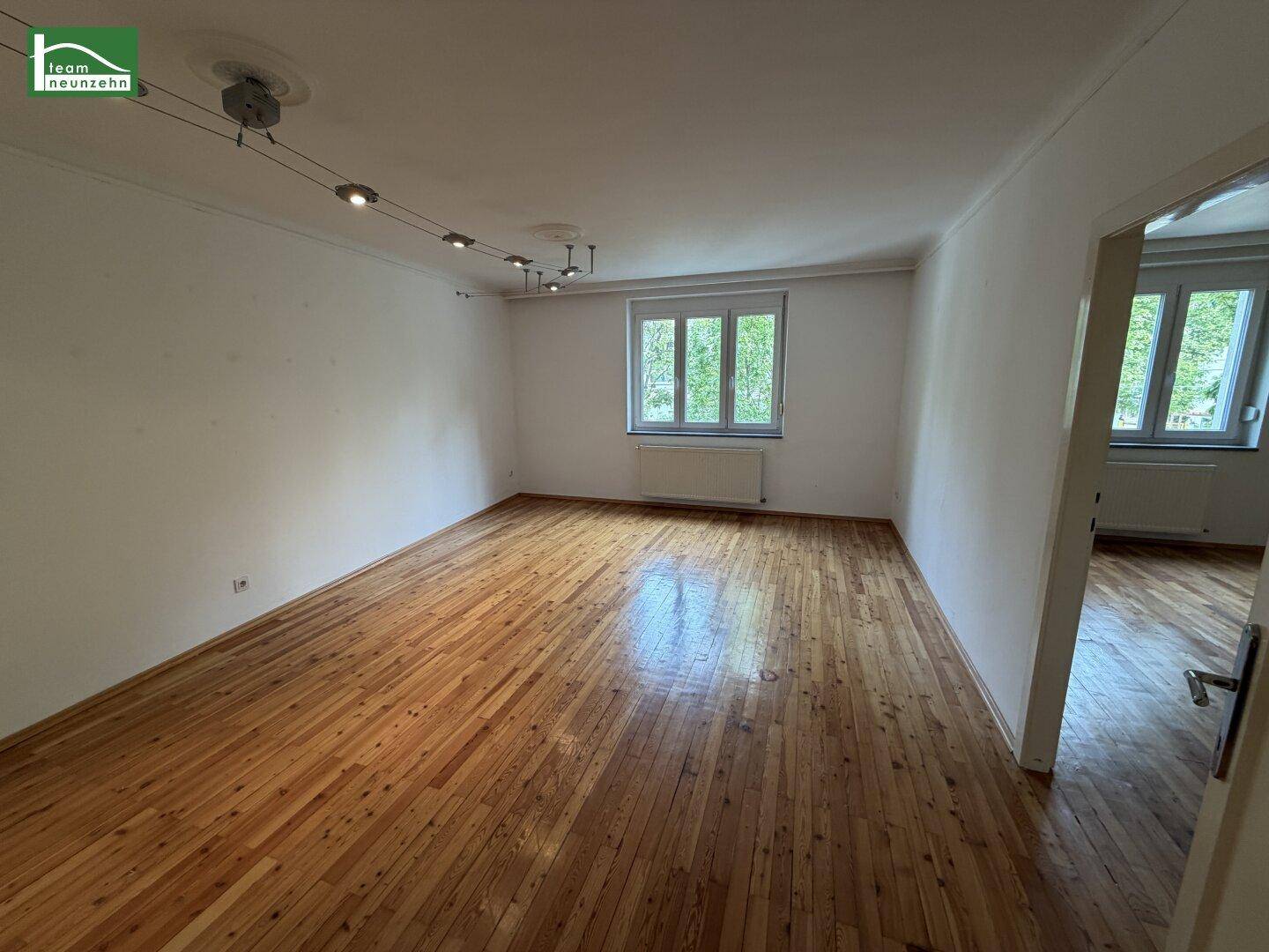 Geräumiges, leeres Zimmer mit hellem Holzparkettboden und Blick auf grüne Bäume.