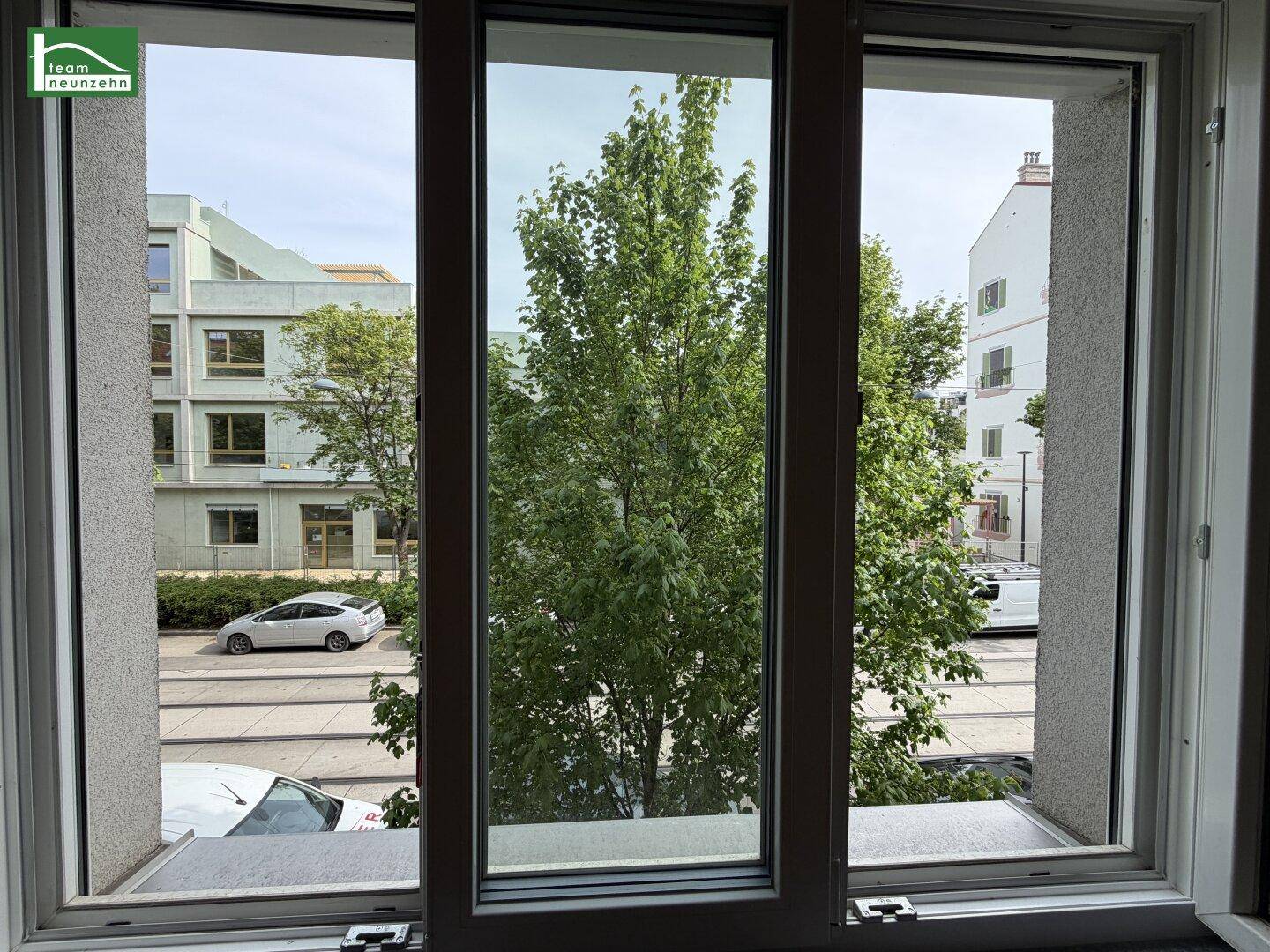 Blick aus dem Fenster auf die belebte Straße mit Bäumen und gegenüberliegenden Gebäuden.