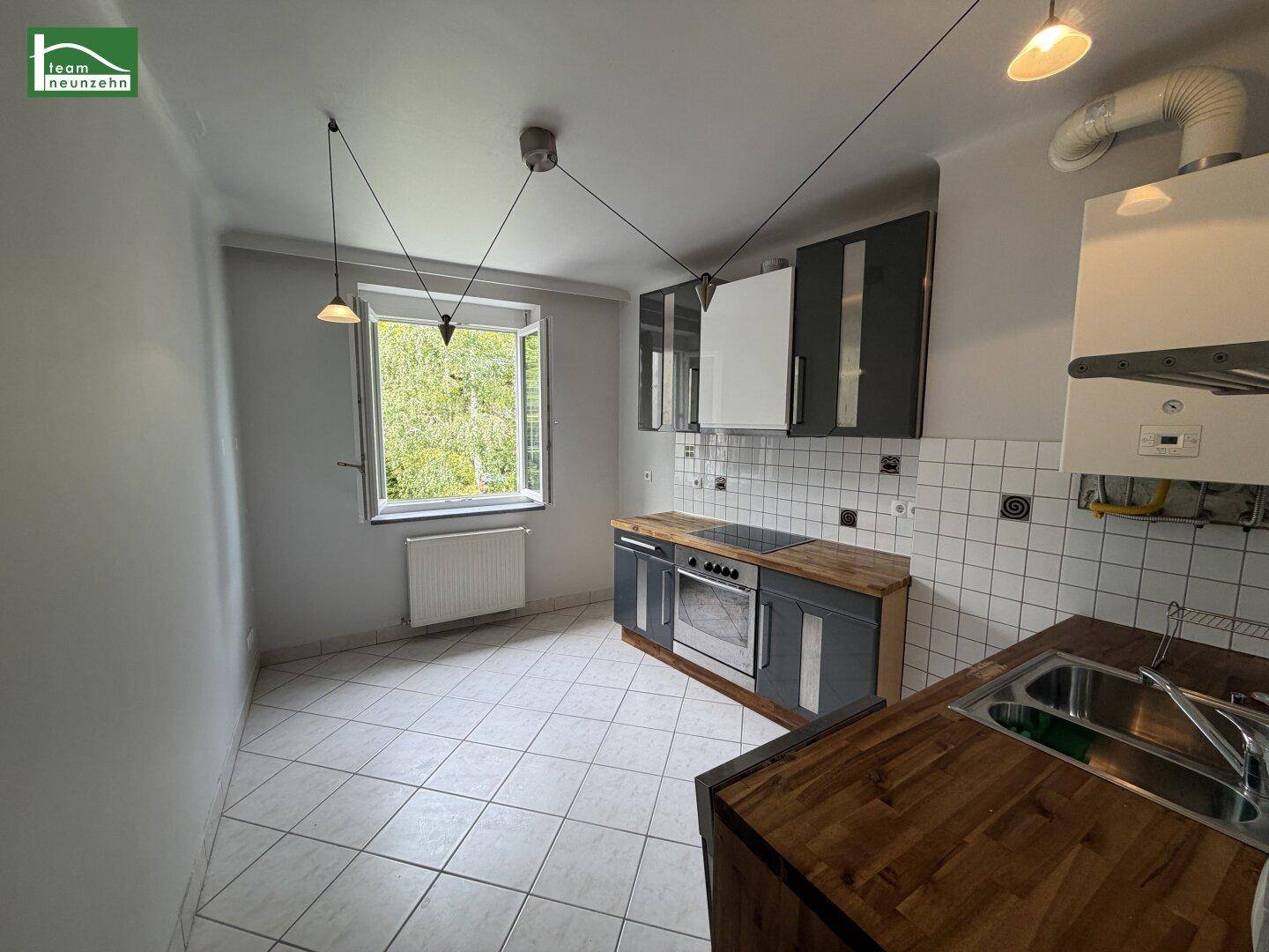 Funktionale Küche mit Holz-Arbeitsplatten, Fliesenboden und einem Fenster zum grünen Hof.