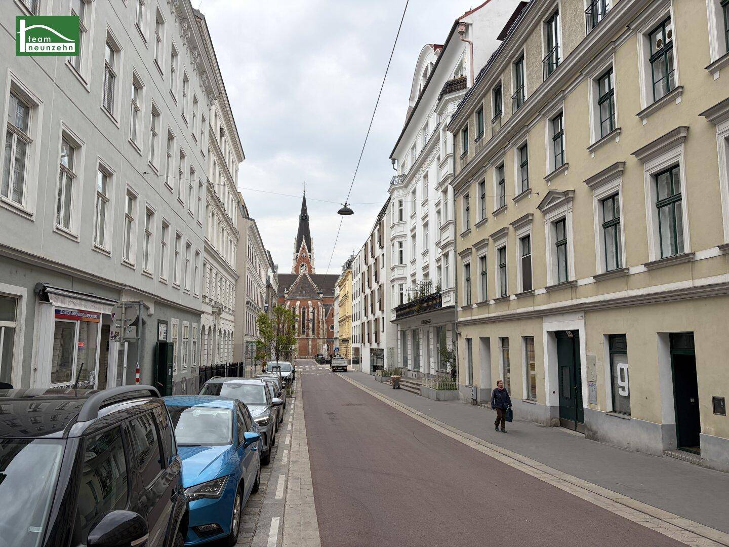 Historische Straße mit parkenden Autos und einem Kirchturm im Hintergrund.