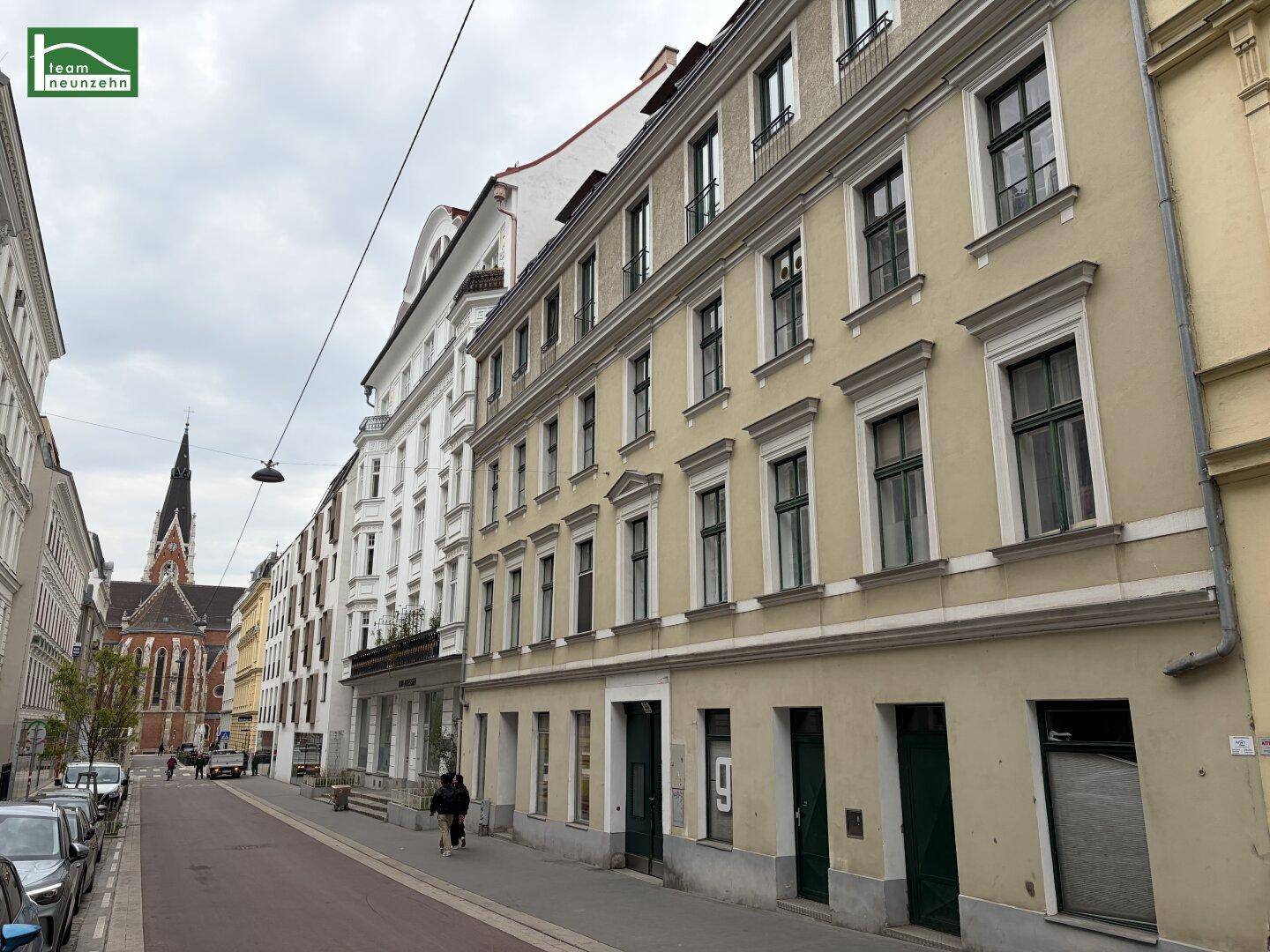 Historische Straße mit parkenden Autos und einem Kirchturm im Hintergrund.