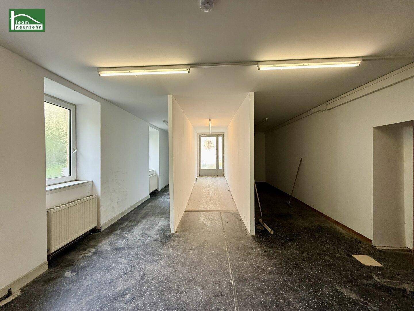 Langer Flur mit Betonboden, einfachen Leuchtstoffröhren und Fenstern, die Tageslicht hereinlassen.