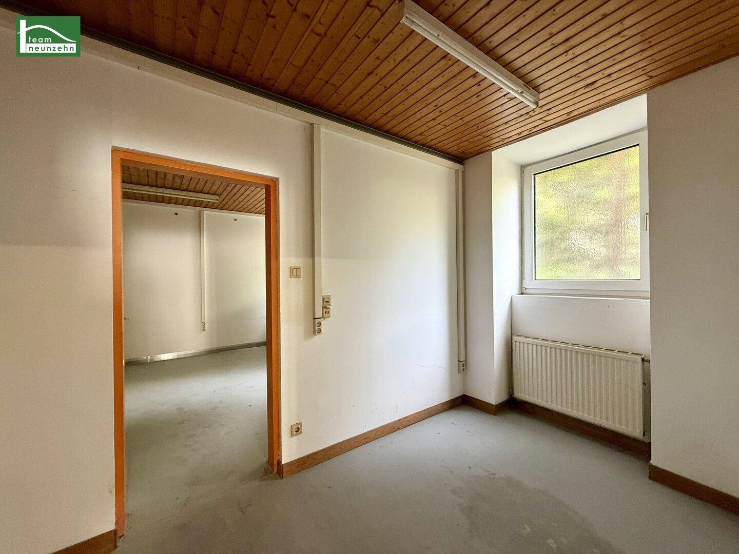 Heller Raum mit Holzdecke, Fenster und Zugang zu einem weiteren Bereich, ideal für Büro oder Lager.