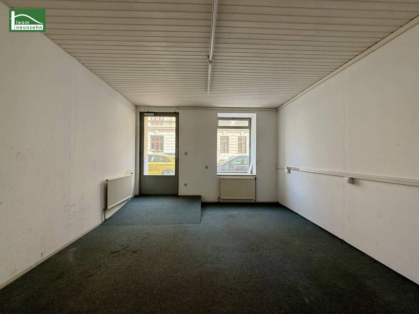 Geräumiger Gewerberaum mit Teppichboden, großen Fenstern zur Straße und einfacher Deckenverkleidung.