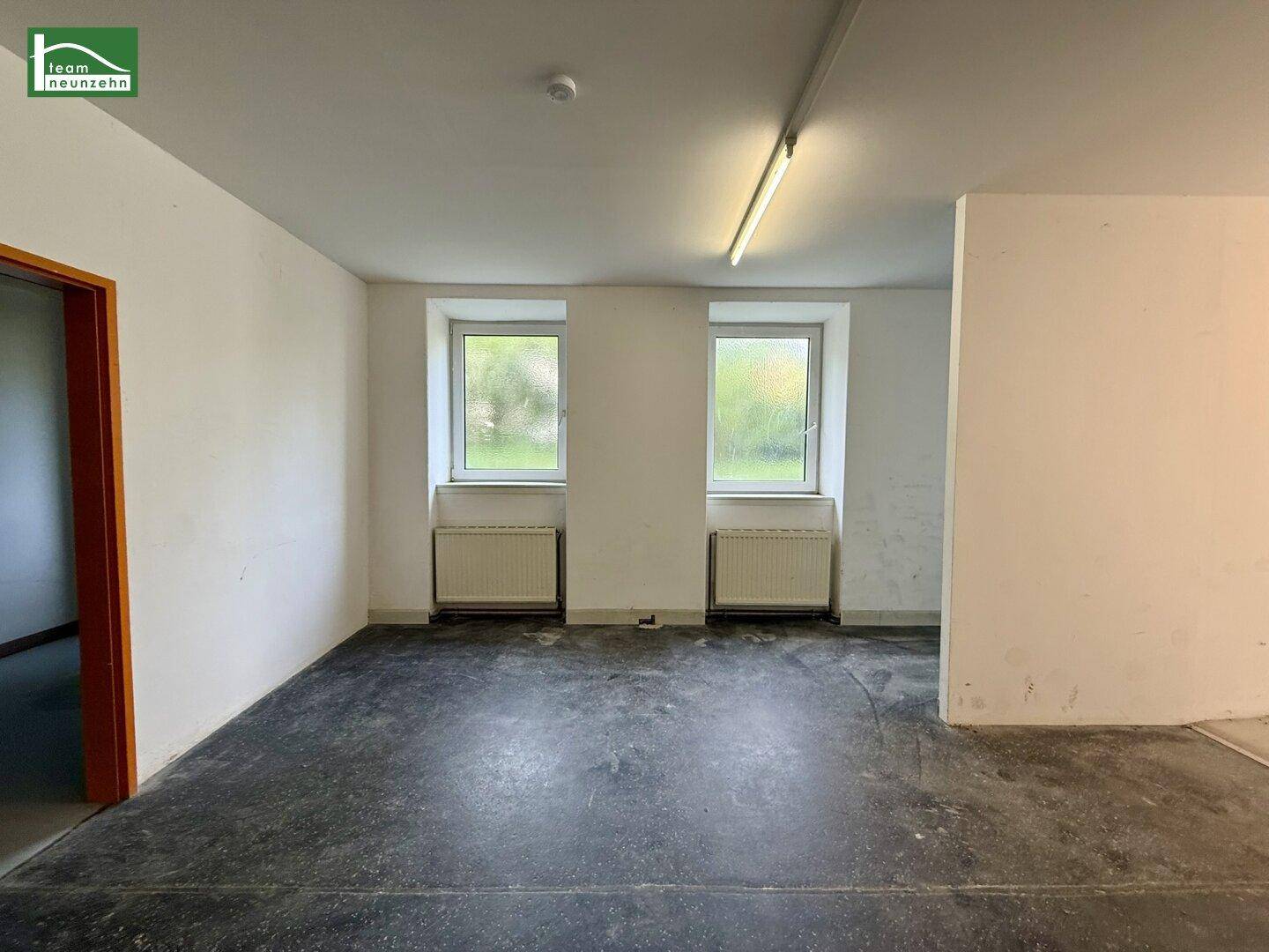 Geräumiger, leerer Raum mit zwei Fenstern, Betonboden und einfacher Beleuchtung, ideal für gewerbliche Nutzung.