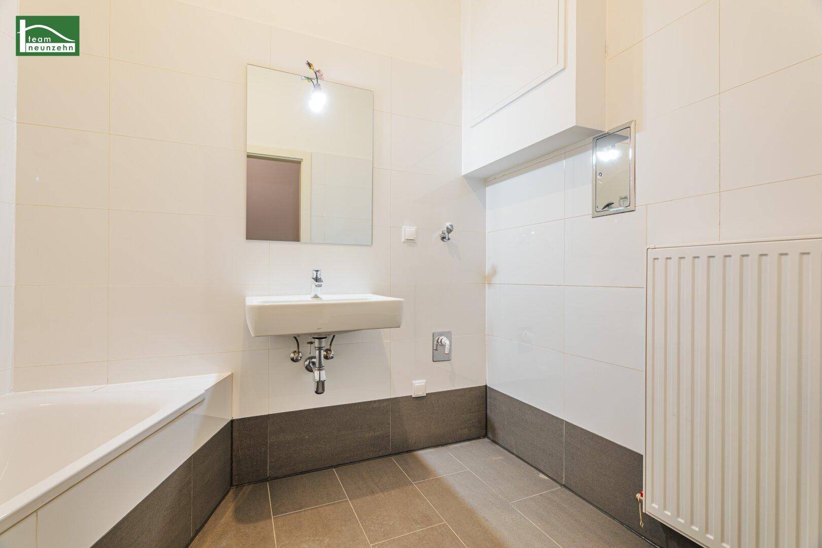Helles Badezimmer mit Waschbecken, Spiegel und Heizkörper, gefliest in Weiß und Grau.