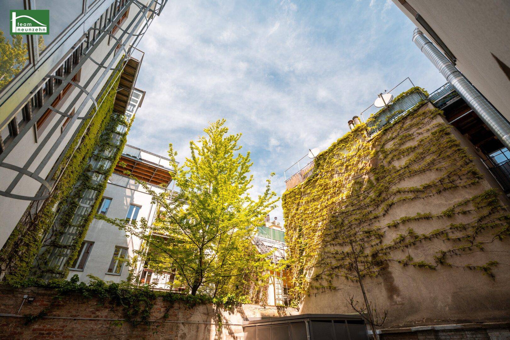 Grüner Innenhof zwischen Gebäuden mit bewachsenen Wänden und Bäumen unter blauem Himmel.