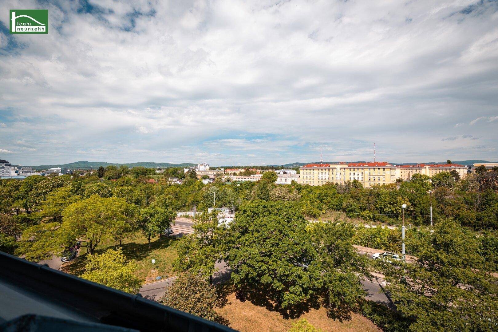 Weitläufiger Ausblick über grüne Bäume und die Stadtlandschaft unter blauem Himmel.