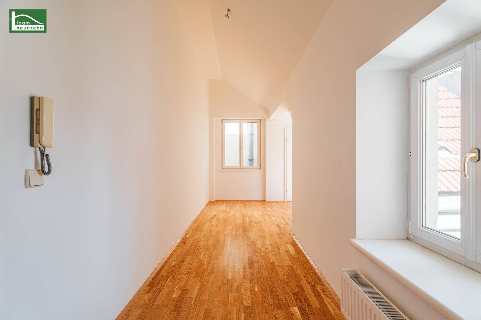 Langer, heller Flur mit Holzboden, Fenstern und einer Gegensprechanlage an der Wand.