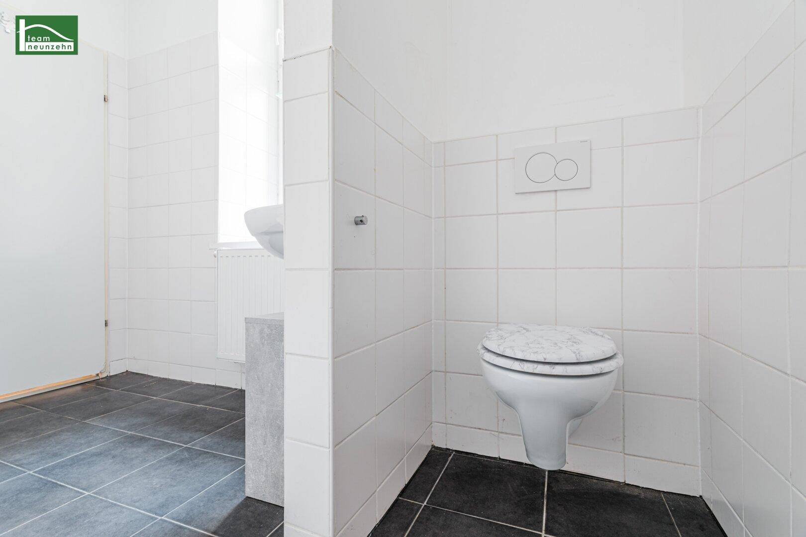 Separates WC mit wandhängender Toilette, weißen Fliesen an den Wänden und dunklem Fliesenboden.