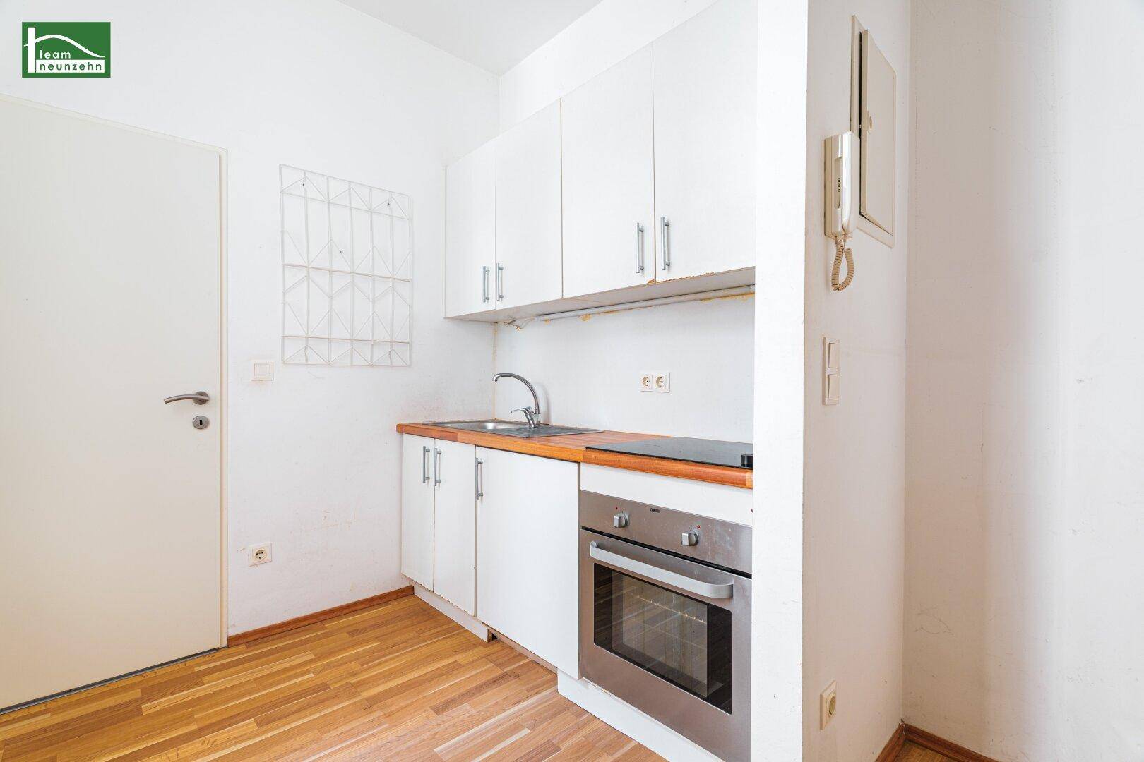 Kleine Einbauküche mit weißen Schränken, Spüle, Backofen und Kochfeld auf Holzfußboden.