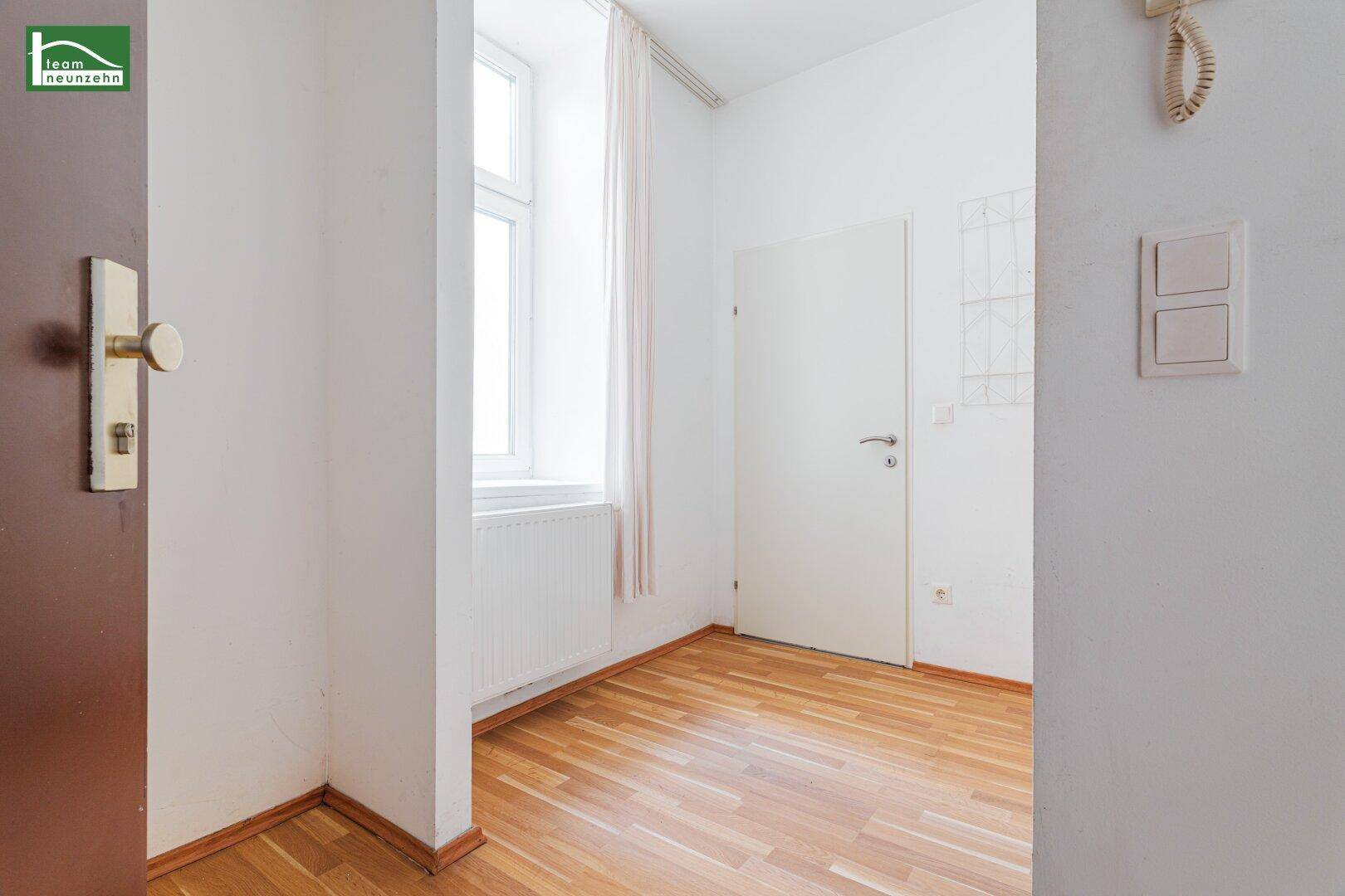 Ecke eines hellen Zimmers mit Fenster, Heizkörper und einer Tür, ausgestattet mit Holzfußboden.