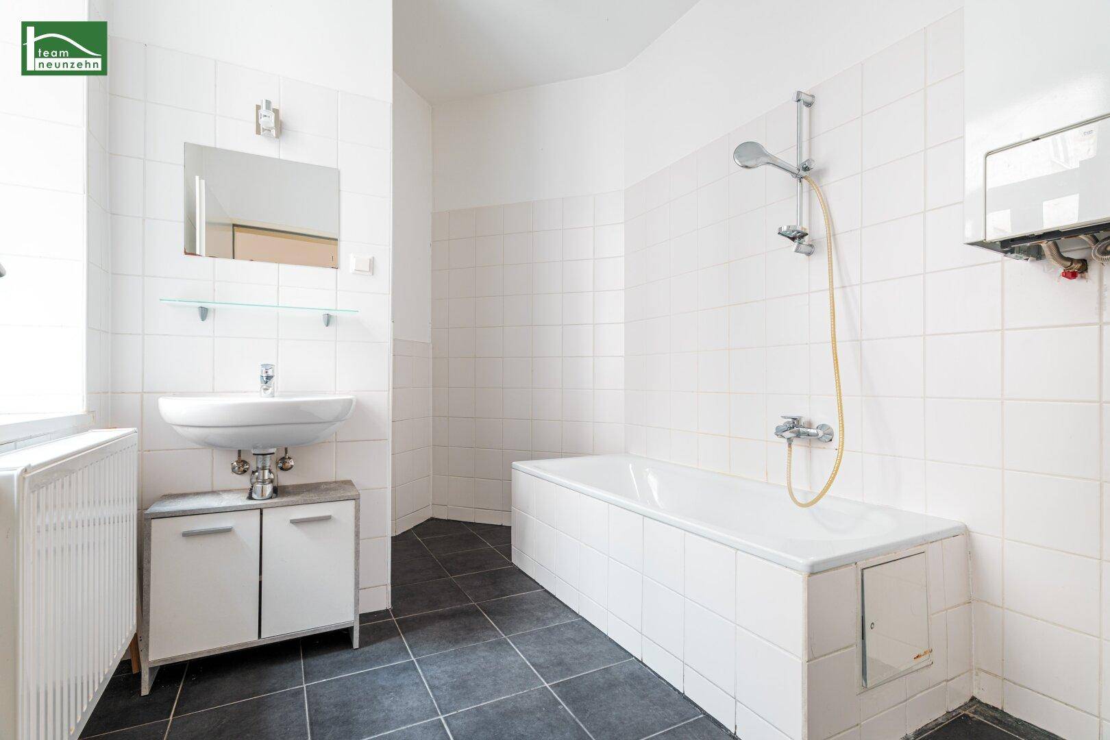 Badezimmer mit weißen Wandfliesen, dunklem Boden, Badewanne mit Duschfunktion und Waschbecken.