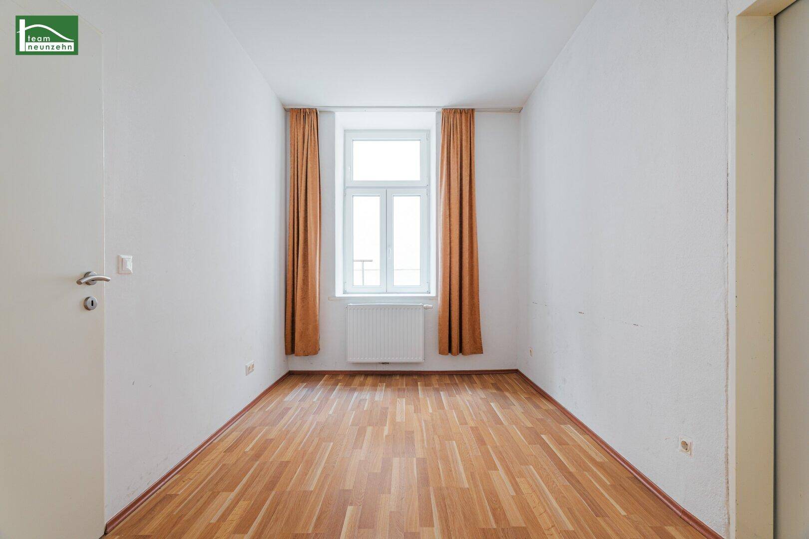 Helles, leeres Zimmer mit Holzfußboden, Fenster und orangefarbenen Vorhängen.