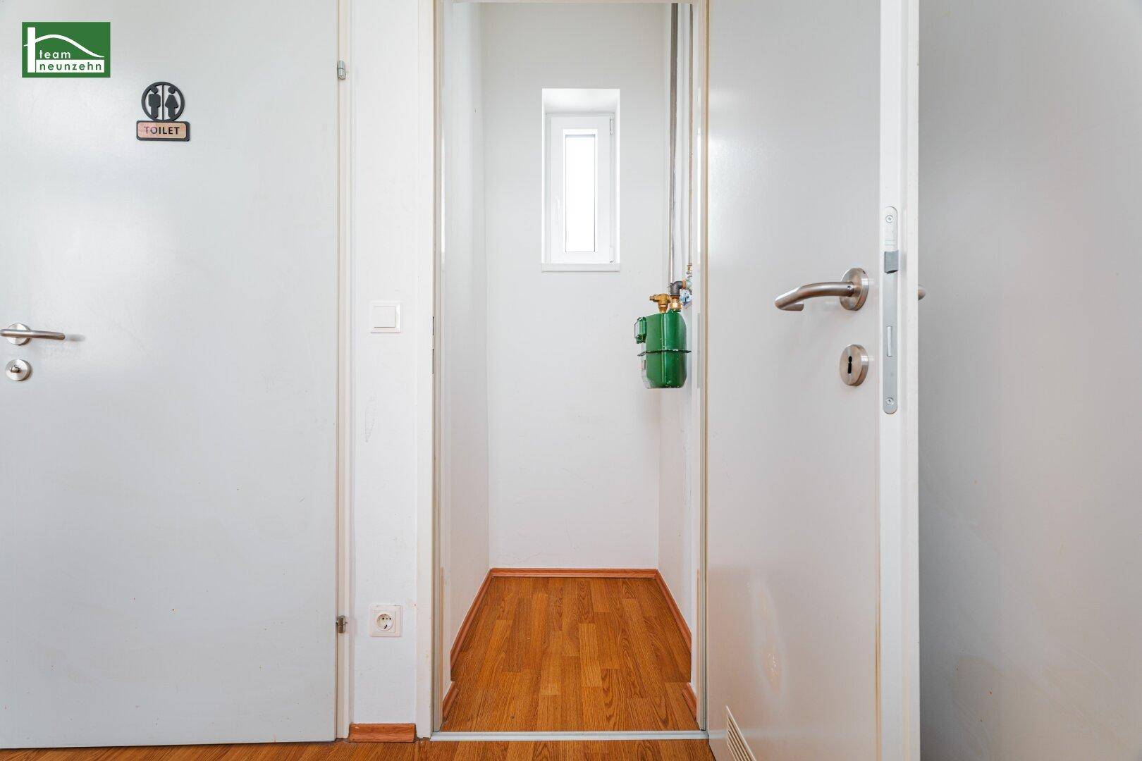 Flur mit Holzboden, der zu einem kleinen Raum mit Fenster führt, daneben eine Toilette.