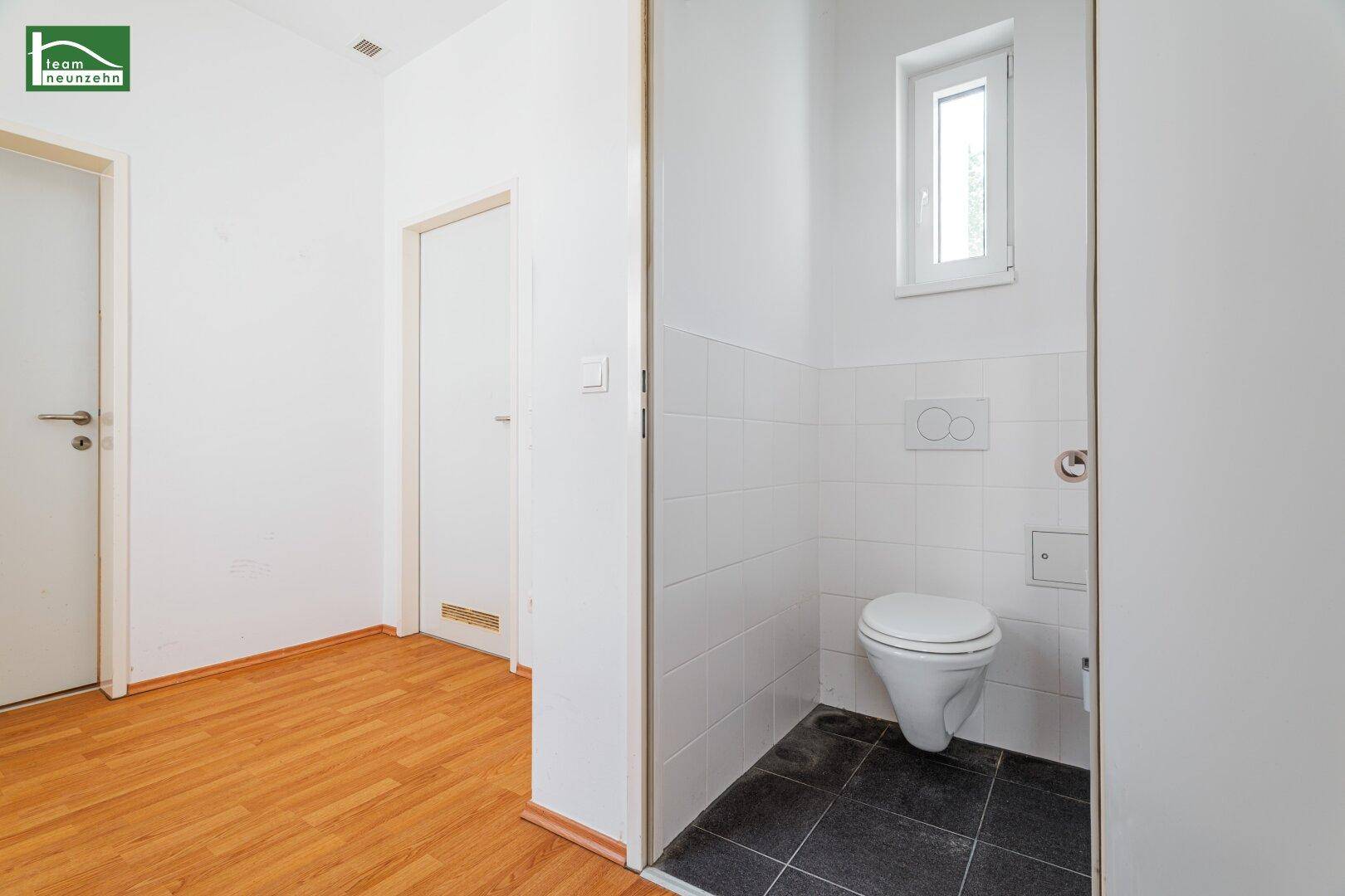 Separate Toilette mit wandhängendem WC, kleinem Fenster und dunklem Fliesenboden.