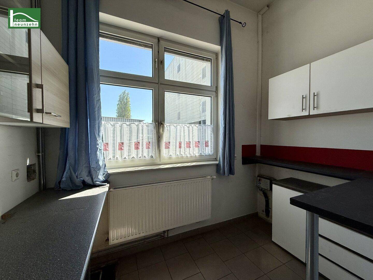 Kleine Küche mit Fenster, blauen Vorhängen, Arbeitsplatten und weißen Hängeschränken.