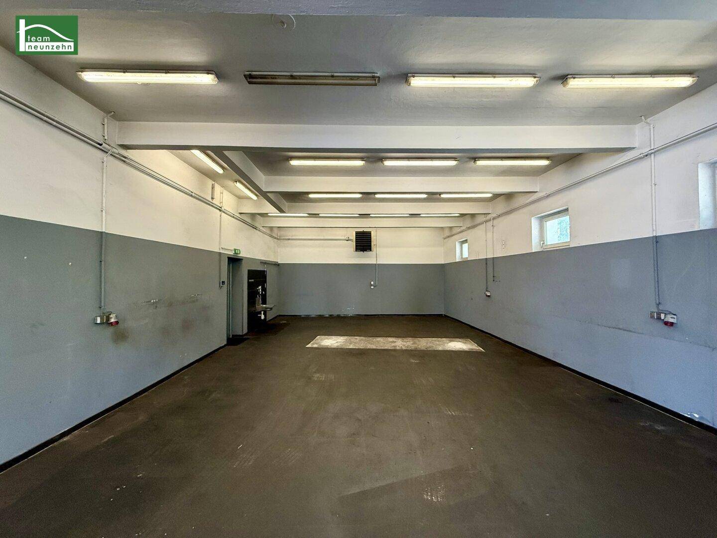 Lange Werkstatthalle mit grauen Wänden, Betonboden und Reihen von Deckenleuchten.