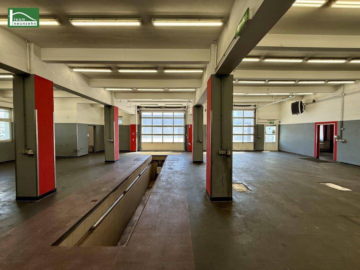 Werkstatthalle mit Fahrzeuggruben, roten Stützpfeilern und großen Garagentoren für Fahrzeuge.