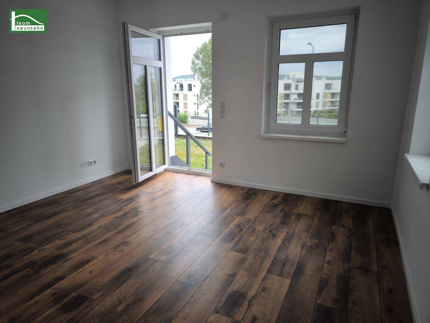 Heller, leerer Raum mit Laminatboden und Zugang zu einem Außenbereich mit Blick auf Gebäude.