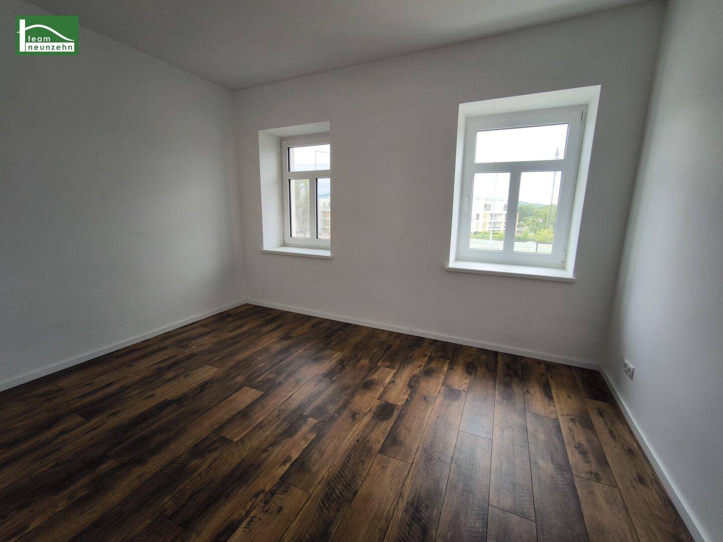 Leerer Raum mit Laminatboden und zwei Fenstern, die für natürliches Licht sorgen.