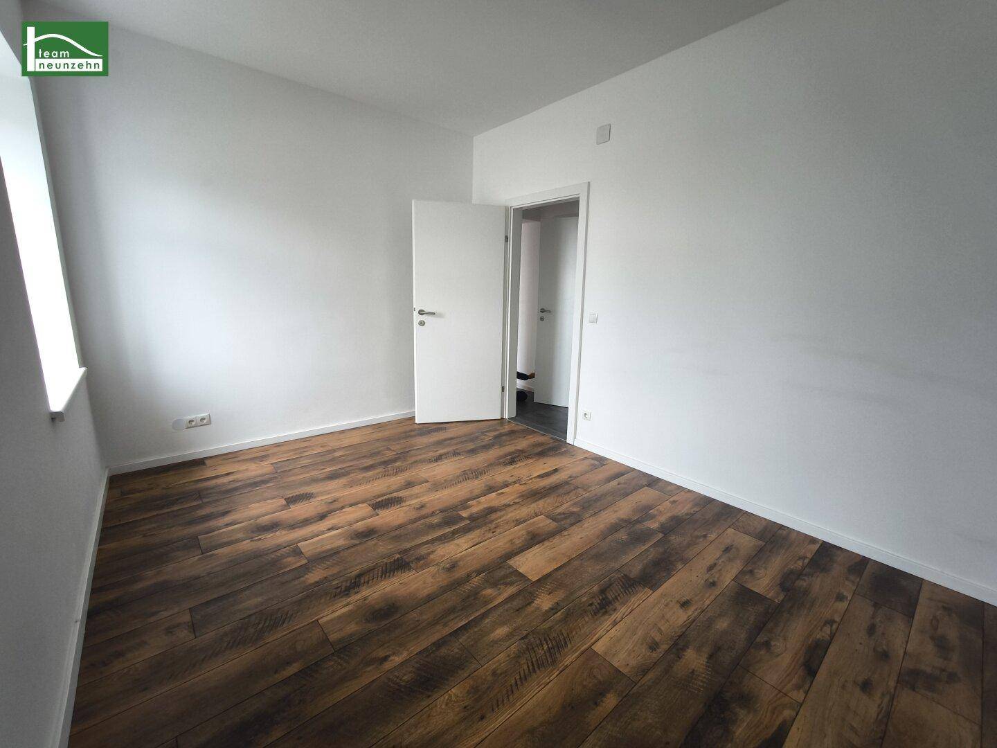 Leerer Raum mit Laminatboden und offener Tür, die den Zugang zu einem weiteren Zimmer ermöglicht.