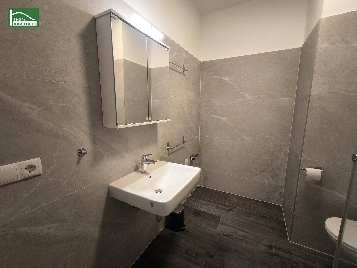 Modernes Badezimmer mit grauen Fliesen, Waschbecken, Spiegelschrank und Toilette.