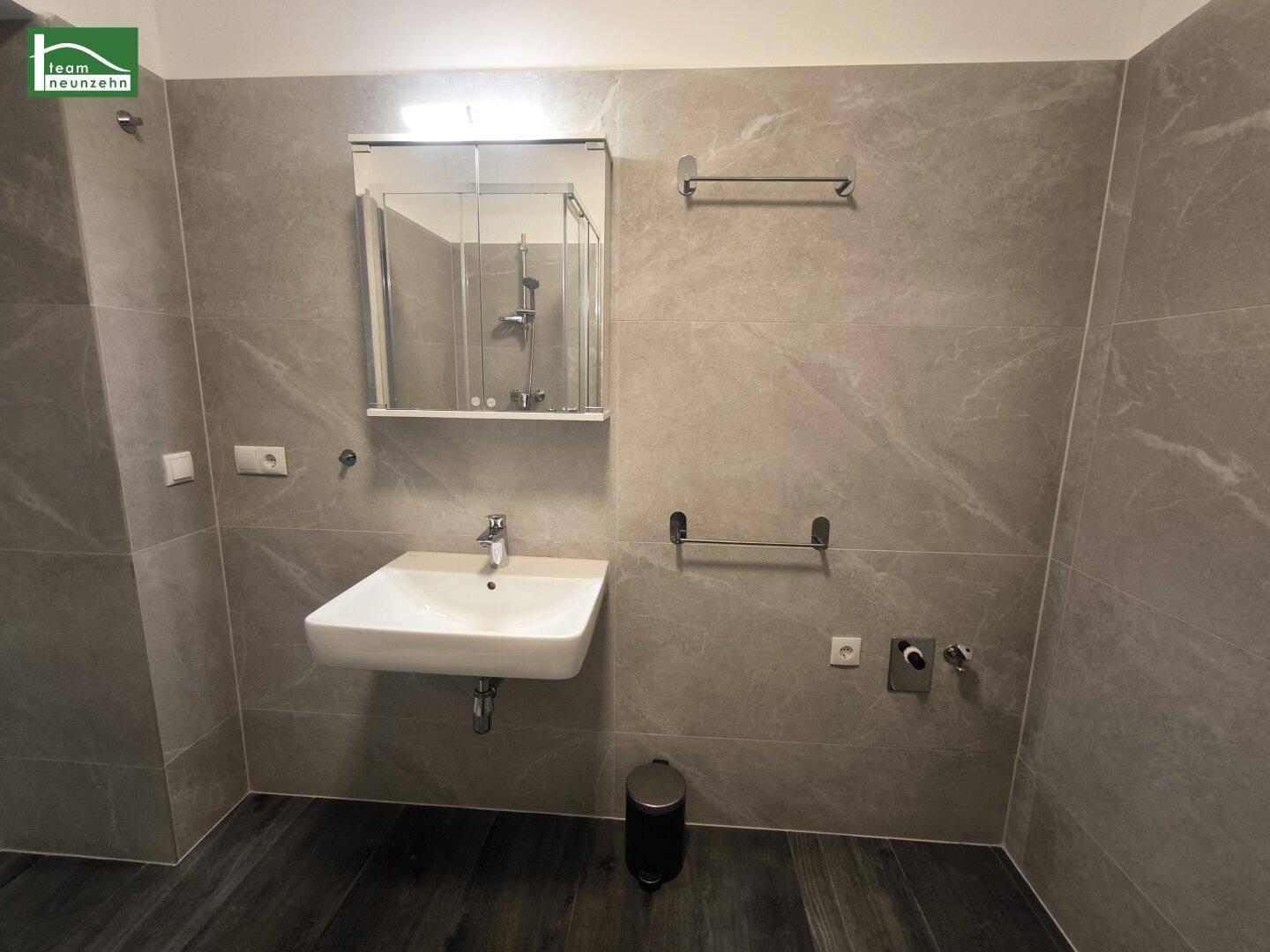 Modernes Badezimmer mit grauen Fliesen, Waschbecken und einem Spiegel, der die Dusche reflektiert.