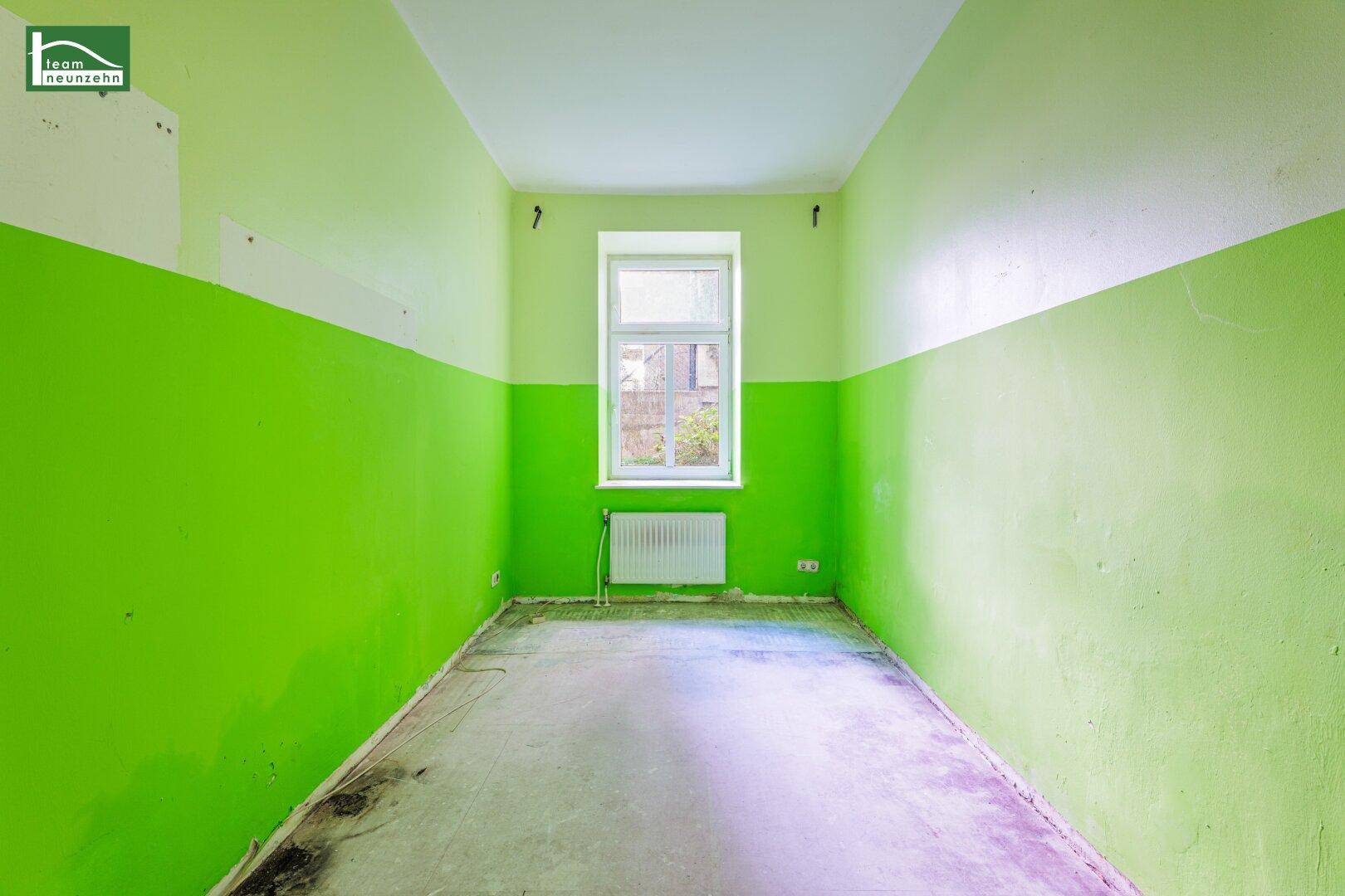 Leerer Raum mit auffällig grün-weißen Wänden, einem Fenster und einem Heizkörper.