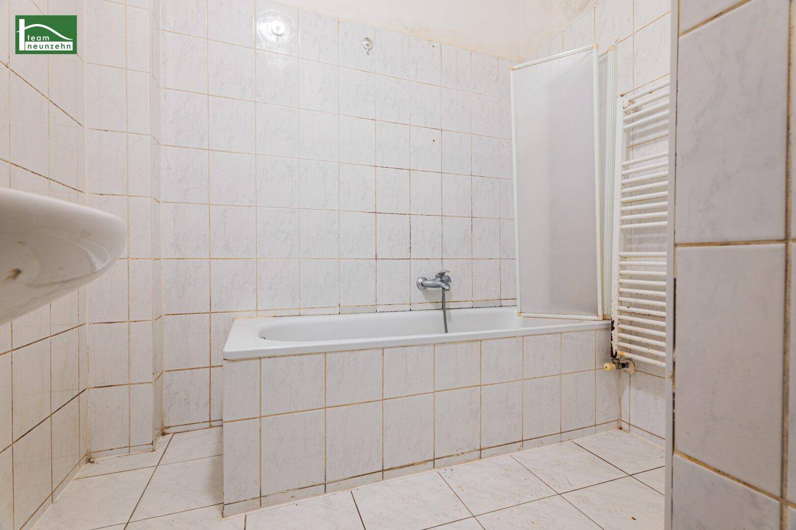 Badezimmer mit weißen Fliesen, Badewanne mit Duschwand und einem Handtuchheizkörper.