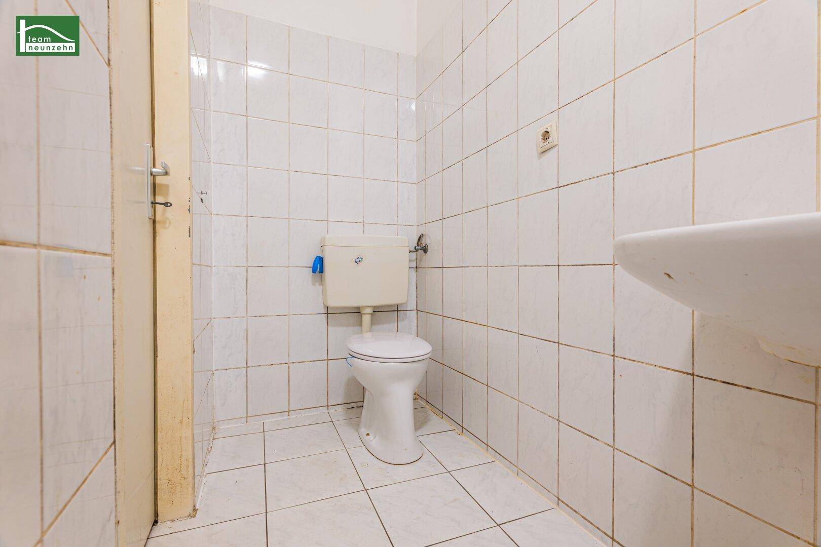 Separate Toilette mit weißen Fliesen, WC und einem kleinen Waschbecken.