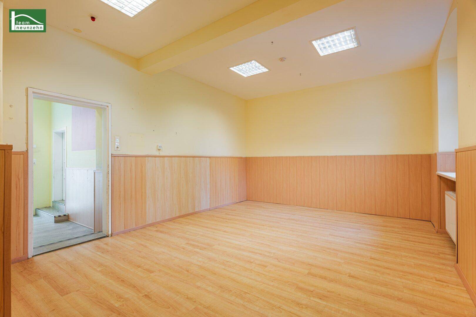 Geräumiger, heller Raum mit Laminatboden und Holzpaneelen, mit Zugang zu einem Nebenraum.
