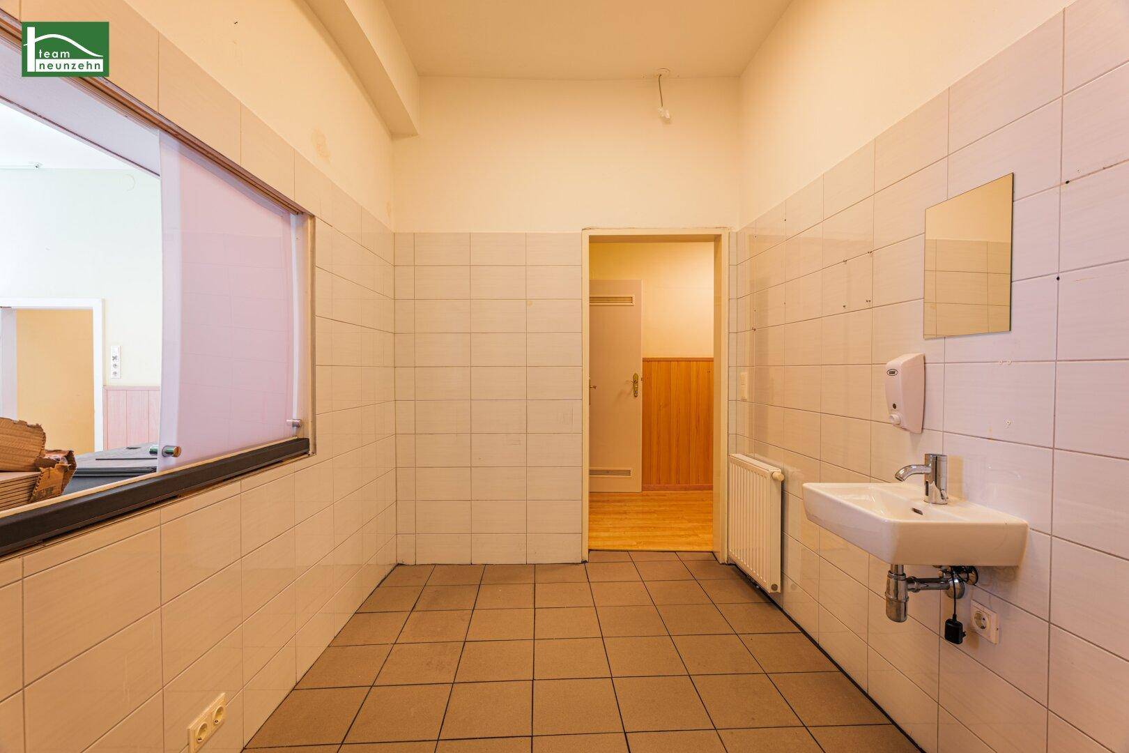 Heller, gefliester Waschraum mit Waschbecken und Fenster, führt zu einem weiteren Bereich.