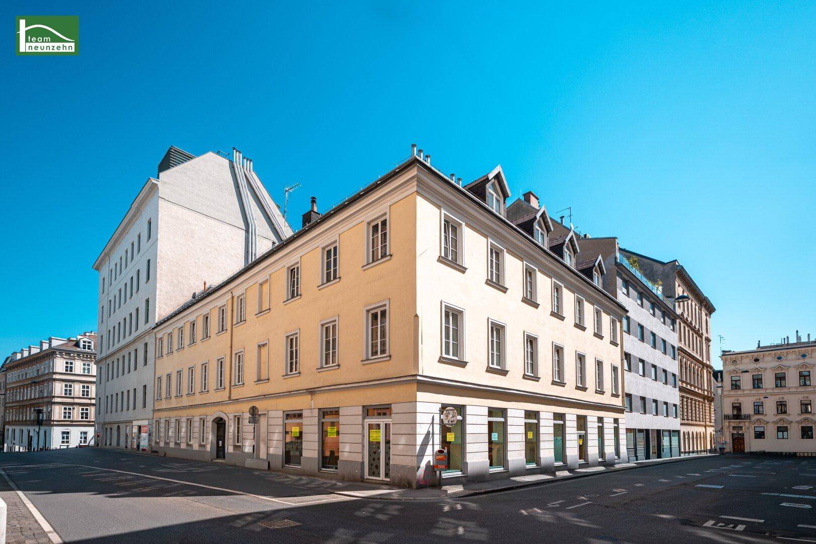 Historisches Eckgebäude mit mehreren Etagen und Geschäften im Erdgeschoss an einer sonnigen Straße.