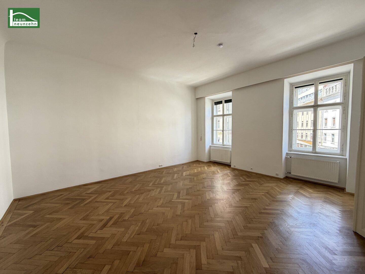 Großes, leeres Zimmer mit Fischgrätparkett und zwei hohen Fenstern, vielseitig nutzbar.