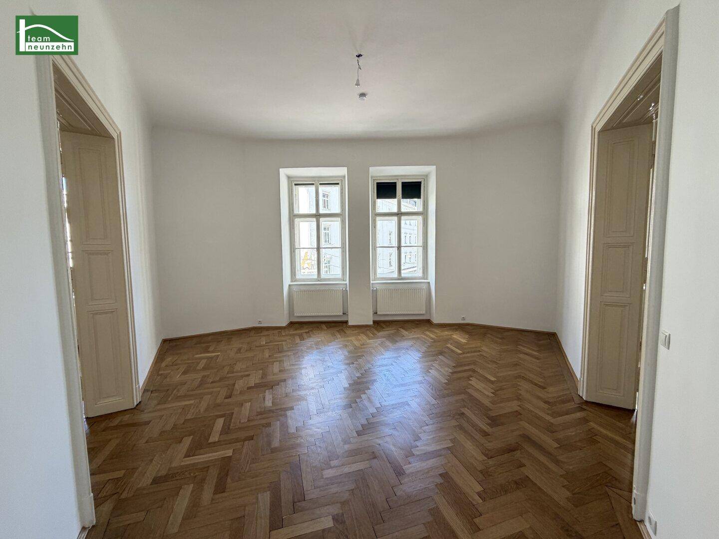 Helles, leeres Zimmer mit Fischgrätparkett und zwei hohen Fenstern, bereit zur Einrichtung.