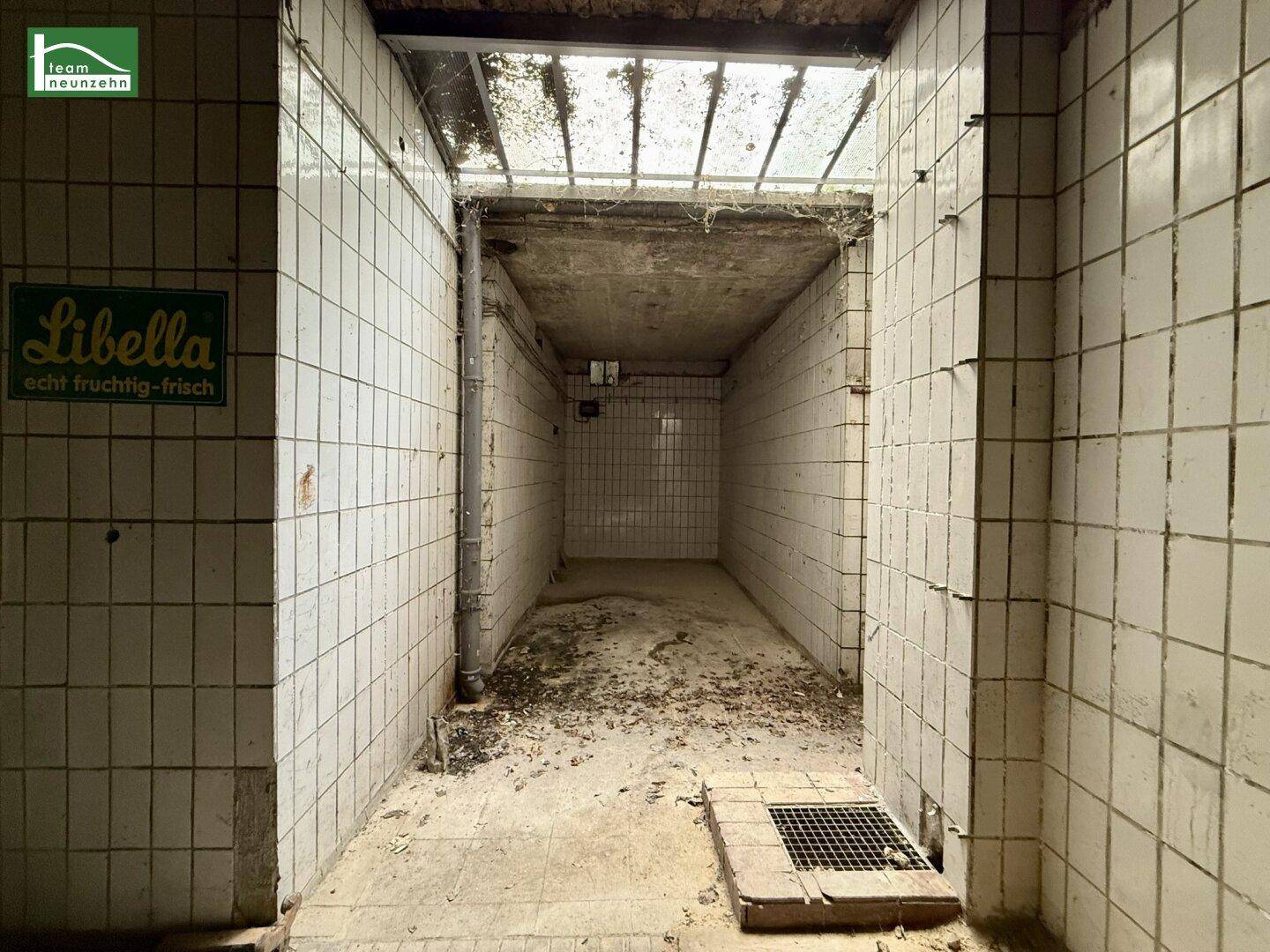 Alter, gefliester Kellergang mit Oberlicht und Abfluss, in sanierungsbedürftigem Zustand.