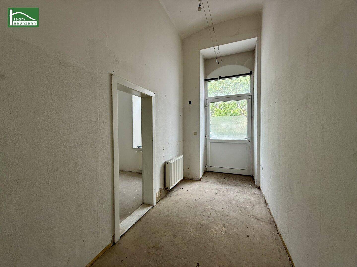 Schmaler Flur mit hoher Tür und Fenster, weißen Wänden und Betonboden, in Renovierung.