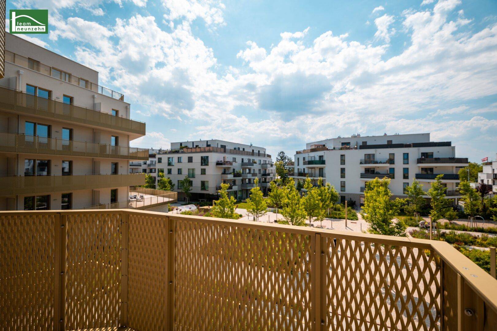 Weiter Blick vom Balkon über die grüne Umgebung und die modernen Wohngebäude unter blauem Himmel.