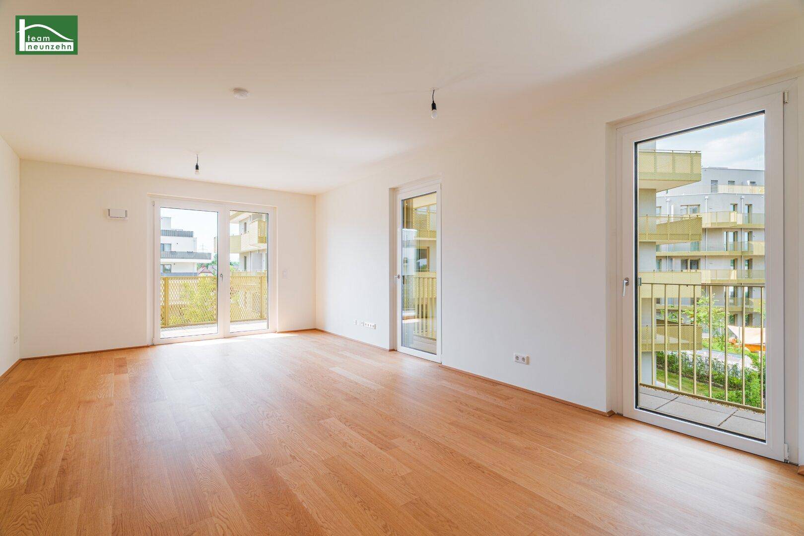 Geräumiges, helles Zimmer mit Holzboden und mehreren Balkonzugängen in einem Neubau.