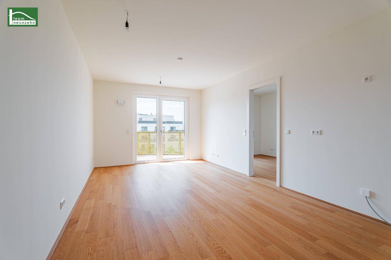 Helles, leeres Wohnzimmer mit Holzboden und Zugang zu einem Balkon.