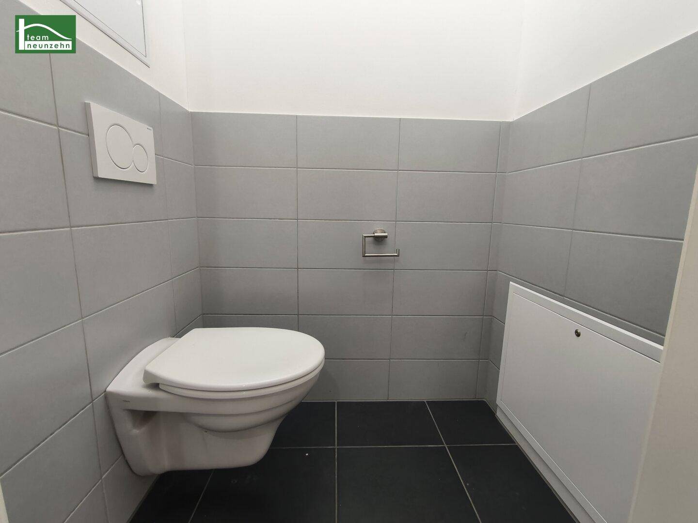 Separates WC mit wandmontierter Toilette und grauen Fliesen an den Wänden.