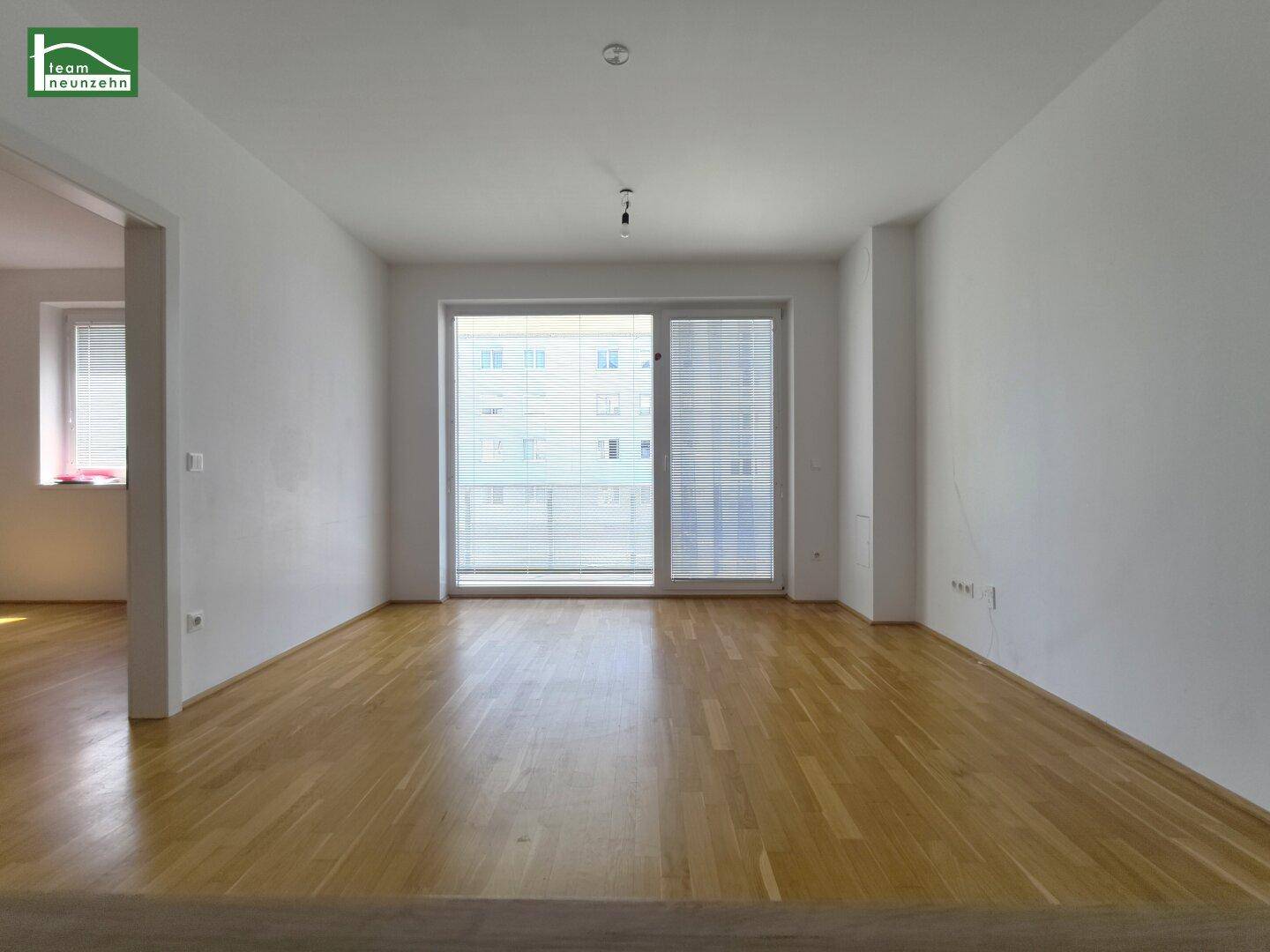 Helles, leeres Wohnzimmer mit Parkettboden und großem Fenster mit Zugang zum Balkon.
